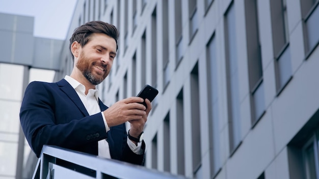 Stylowy przystojny biznesmen trzymający telefon czytający e-mail stoi na ulicy uśmiechnięty pisze wiadomość