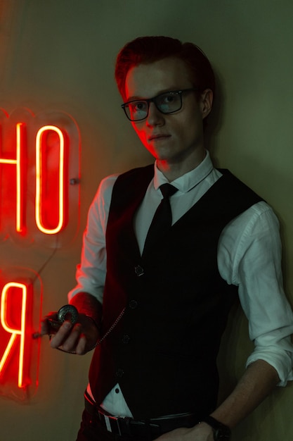 Stylowy przystojny młody biznesowy model człowieka w okularach w modnych ubraniach wizytowych z koszulą, kamizelką i krawatem z zegarem w pobliżu zielonej ściany z czerwonym światłem neonowym. Czas, praca i biuro