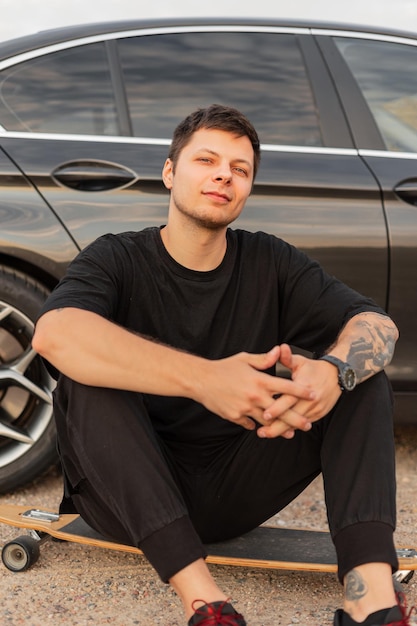 Stylowy przystojny młody hipster facet w czarnych ubraniach z tatuażem na ramieniu, siedząc na longboard w pobliżu jego samochodu Odpoczynek i letni weekend