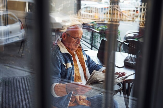 Zdjęcie stylowy senior w modnych ubraniach i okularach siedzi w kawiarni i czyta gazetę.