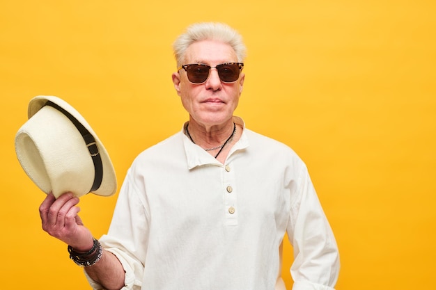 Stylowy starszy mężczyzna w okularach przeciwsłonecznych i białej koszuli trzymający kapelusz panama