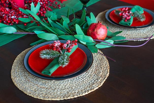 Stylowy świąteczny wzór z zieloną serwetką ozdobioną gałązką z jagodami i gałązką jodły, czerwone talerze na drewnianym stole. Boże Narodzenie lub koncepcja nowego roku. Nakrycie stołu na świąteczny posiłek.