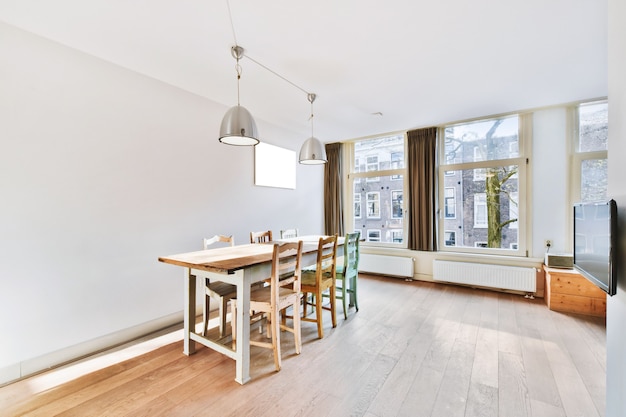 Stylowy wystrój domu ze strefą jadalną z lampami wiszącymi nad drewnianym stołem i krzesłami w nowoczesnym mieszkaniu z dużymi oknami i minimalistycznym designem