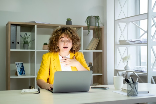 Sukces w biznesie Zaskoczony pracownik Dobre wieści Ciało pozytywne Podekscytowany szczęśliwy kręcone włosy z nadwagą Kobieta pracownik przy laptopie pracujący online w jasnym wnętrzu miejsca pracy