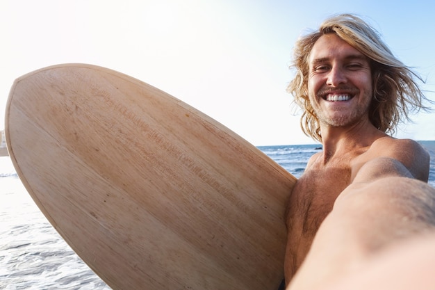 Surfer robi selfie na plaży po sesji surfowania, trzymając deskę z drewna w stylu vintage - Skup się na twarzy