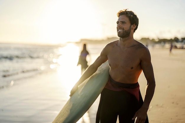 Surfer w strojach kąpielowych z deską surfingową stoi na plaży i patrzy na wodę