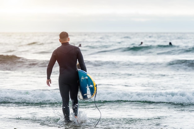 Surfer wchodzący do morza z deską surfingową patrzący na innych surferów