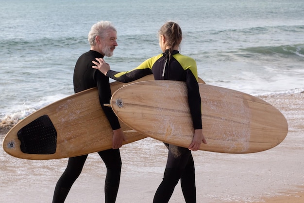 Surferów płci męskiej i żeńskiej, korzystających z wakacji razem. Szczęśliwy mężczyzna i kobieta w kombinezonach spacerujących wzdłuż brzegu morza z deskami, patrzący na siebie z miłością. Koncepcja sportu, aktywności rekreacyjnej osób starszych