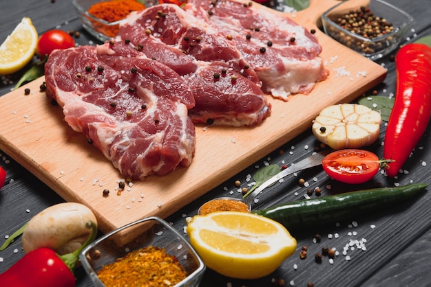 Surowe mięso wieprzowe na drewnianej desce do krojenia przy kuchennym stole do gotowania steku wieprzowego pieczonego lub grillowanego ze składnikami ziół i przypraw, Świeża wieprzowina
