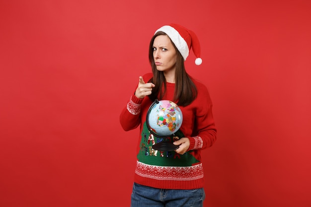 Surowe młoda dziewczyna Santa w Boże Narodzenie kapelusz trzymając świat świata wskazując palcem wskazującym na aparat na białym tle na tle czerwonej ściany. Szczęśliwego nowego roku 2019 celebracja party wakacje koncepcja. Makieta miejsca na kopię.