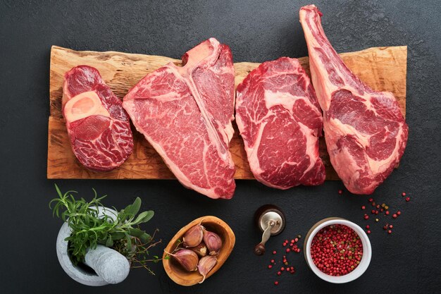 Surowe steki Prime Różnorodność świeżych steków mięsnych Black Angus Prime Tbone New York Ribeye Striploin Tomahawk deska do krojenia na czarnym lub ciemnym tle Zestaw różnych klasycznych steków Widok z góry