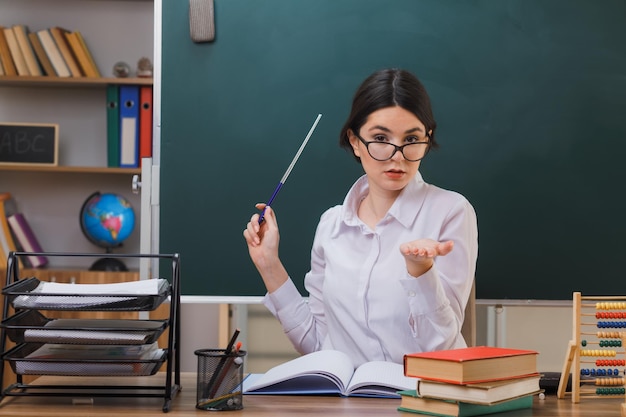 Surowe Wyciąganie Ręki Do Kamery Młoda Nauczycielka W Okularach Wskazuje Na Tablicę Ze Wskaźnikiem Siedzącym Przy Biurku Z Szkolnymi Narzędziami W Klasie