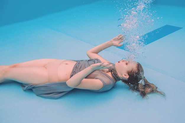 surrealistyczny portret młodej kobiety w szarej sukience i szaliku z koralików pod wodą w basenie
