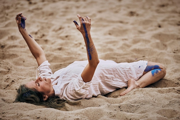 Svetlogorsk Rosja 08152021 Kobiecy spektakl taneczny na piaszczystej plaży Tancerka leżąca na plecach na piasku i poruszająca rękami na świeżym powietrzu występ artystyczny na świeżym powietrzu Ekspresyjny powolny taniec