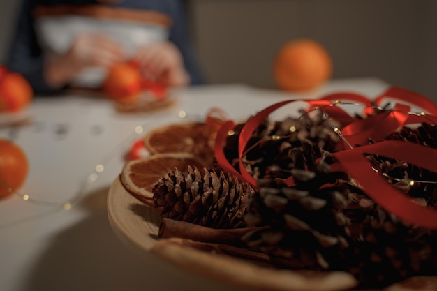 Świąteczna dekoracja z szyszek sosnowych, lekkiej girlandy i czerwonej wstążki na drewnianym talerzu na stole w for