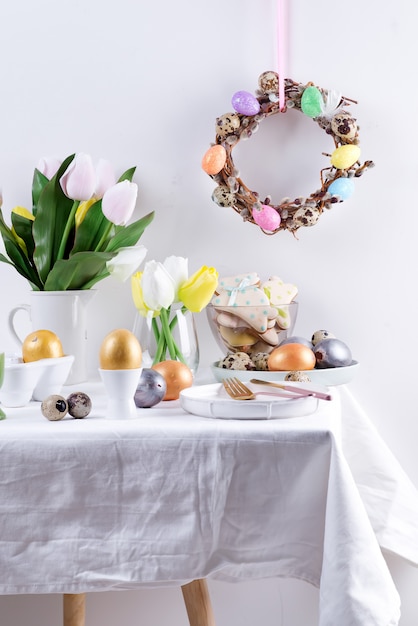 Świąteczna kompozycja gratulacyjna serwowanego stołu z ręcznie malowanymi jajkami, pieczonymi ciastkami, świeżymi wiosennymi kwiatami tulipanów i świątecznym wieńcem na jasnoszarej ścianie. Wesołych Świąt Wielkanocnych.