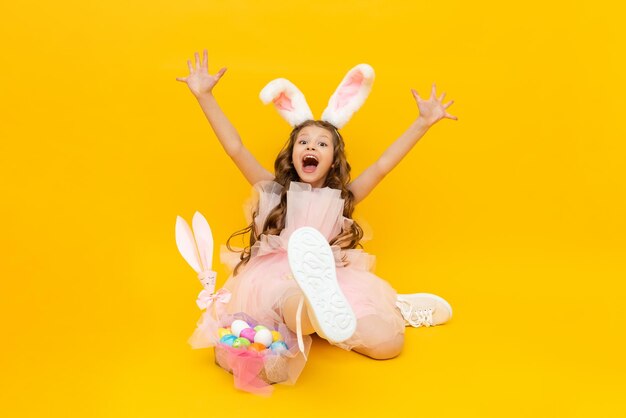 Świąteczna Wielkanoc Mała dziewczynka przedstawia zajączka wielkanocnego z koszem kolorowych jaj Bardzo szczęśliwe dziecko z uszami królika na żółtym odosobnionym tle