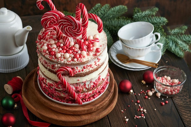 Świąteczne ciasto wielowarstwowe ozdobione czerwonymi i białymi konfetti lolli pops na białym stojaku na ciasto Bombki choinkowe gałęzie jodły na starym drewnianym rustykalnym stole Selektywna ostrość