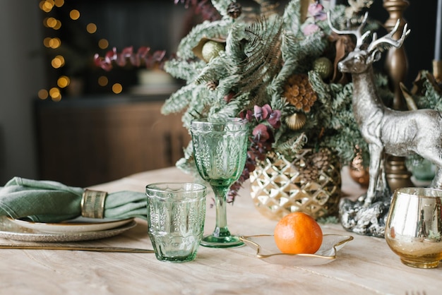 Świąteczne świąteczne ustawienie kolacji Zielone kieliszki do wina gałęzie jodły lub świerku w wazonie talerze sztućce i mandarynka na tle świateł bokeh