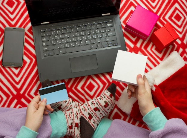 Świąteczne zakupy online. dziewczyna w piżamie i skarpetkach ze świątecznym nadrukiem zamawia prezenty przez Internet za pomocą laptopa. dziewczyna zamawia prezenty przez Internet za pomocą laptopa.