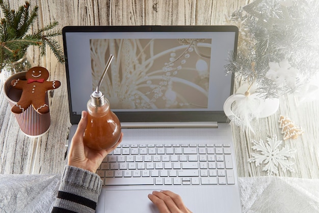 Świąteczny domowy laptop piernik i choinka z dekoracjami Ręce kobiety trzymają żarówkę kubek kakao Koncepcja pracy zdalnej webinarium lub dzwonienie do przyjaciół rodziny z powodu blokady