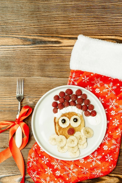 Świąteczny naleśnik w kształcie twarzy świętego mikołaja ze słodką świeżą malinową jagodą, bananem na talerzu, świąteczną skarpetą na drewnianym tle dla dzieci dzieci śniadanie. świąteczny deser spożywczy, dekoracje sylwestrowe