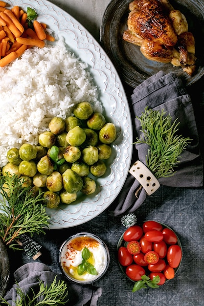 Świąteczny noworoczny stół obiadowy z grillowanym kurczakiem, ryżem i warzywami pieczonymi brukselką, marchewką w ceramicznych miseczkach, dekoracjami świątecznymi na niebieskim lnianym obrusie na szarym tle. Płaski układanie.