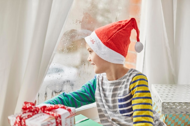 Świąteczny poranek w rodzinnym domu Chłopiec w czapce Mikołaja wyglądający przez okno sypialni