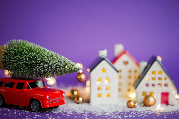 Świąteczny wystrój - czerwony samochód retro na śniegu jedzie obok domów z bajkowymi światłami w choince bokeh z pudełkami prezentowymi na dachu. Zabawka na fioletowym tle. Kartkę z życzeniami nowego roku. Przytulny dom