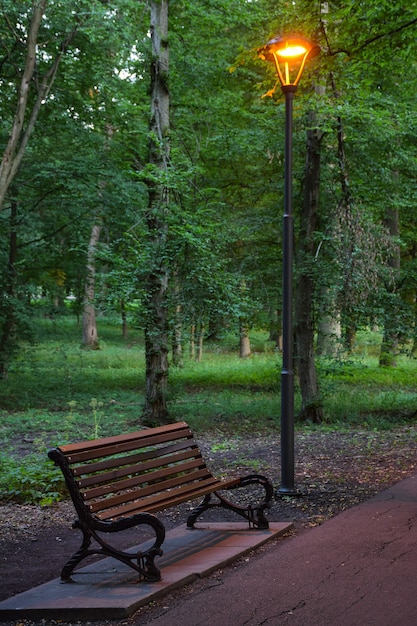 Świecąca latarnia w letnim wieczornym parku, pod ławką do relaksu i chodnikiem