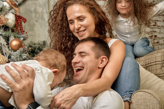 Święta Bożego Narodzenia. Szczęście. Portret Taty, Mamy I Dzieci W Różnym Wieku Siedzą Na Kanapie W Domu Przy Choince I Kominku, Wszyscy Się Uśmiechają. Szczęśliwego Nowego Roku