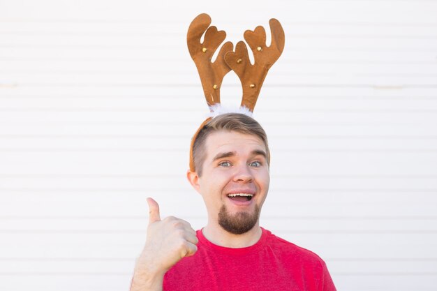 Święta i Boże Narodzenie koncepcja - młody człowiek w rogach jelenia z kciukiem w górę gestem na białym tle.
