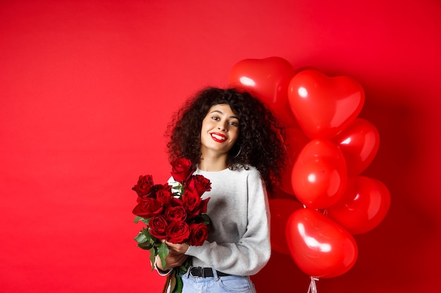Święta I Uroczystości. Szczęśliwa Piękna Kobieta Z Kręconymi Włosami, Otrzymuje Bukiet Róż I Uśmiechnięty, Stojący W Pobliżu Balonów, Czerwone Tło.