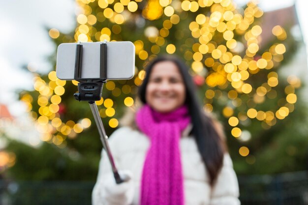 Zdjęcie Święta, technologia i ludzie koncepcja - szczęśliwa kobieta robiąca zdjęcie ze smartfonem selfie stick przy choince