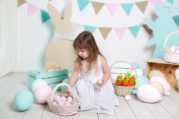 Święta Wielkanocne. Piękna mała dziewczynka w białej sukni składa jaja wielkanocne w koszu. Wiele różnych kolorowych pisanek, kolorowe wnętrze. Zajączek, marchewka i kolorowe flagi. wnętrze i wystrój. rolnik