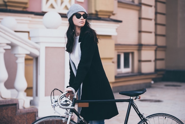Świetny dzień na małą przejażdżkę. Piękna młoda kobieta w okularach przeciwsłonecznych tocząca rower i odwracająca wzrok podczas spaceru na świeżym powietrzu