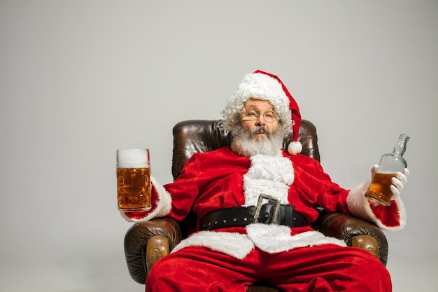 Święty Mikołaj Pije Piwo Siedząc Na Fotelu, Gratulując, Wygląda Na Pijanego I Szczęśliwego. Kaukaski Model Mężczyzna W Tradycyjnym Stroju. Nowy Rok 2020, Prezenty, święta, Zimowy Nastrój. Miejsce Na Reklamę.