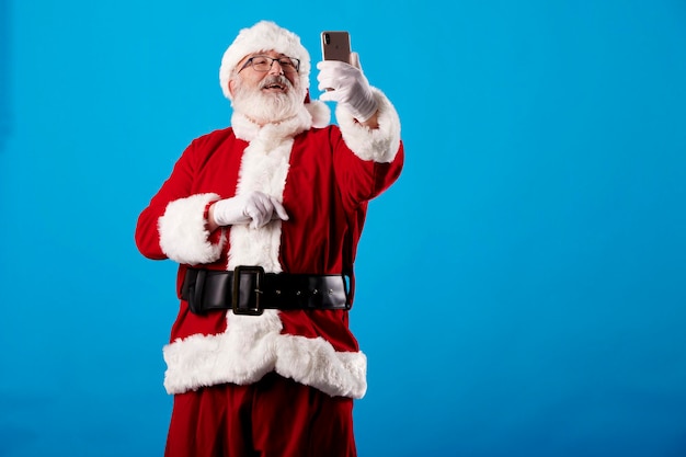 Święty Mikołaj robi selfie telefonem komórkowym na niebieskim tle
