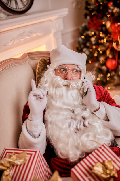 Święty Mikołaj siedzi na kanapie i rozmawia przez telefon komórkowy przy kominku i choinki z prezentami. Nowy rok i Wesołych Świąt, koncepcja wesołych świąt