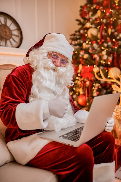 Święty Mikołaj siedzi w swoim domu i czyta e-maile na laptopie z bożonarodzeniowymi prośbami lub listą życzeń przy kominku i drzewie z prezentami. Nowy rok i Wesołych Świąt, koncepcja wesołych świąt