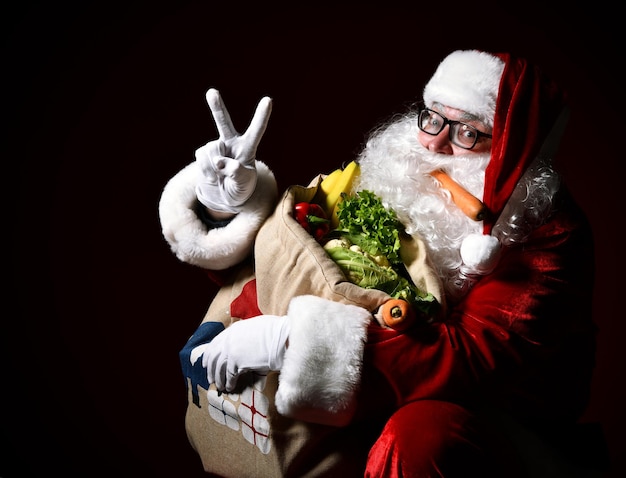 Święty Mikołaj trzyma dużą torbę pełną owoców i warzyw Pokazuje palcami znak pokoju