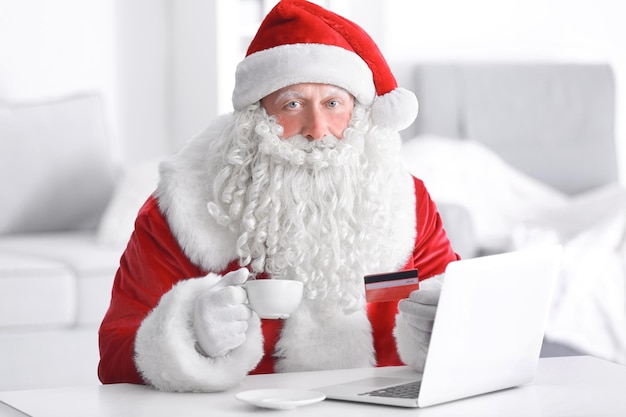 Święty Mikołaj z laptopem i kartą kredytową pije kawę w domu