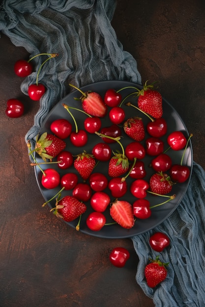 Świeża organiczna letnia mieszanka truskawek i czereśni w okrągłej kamiennej płycie na ciemnej teksturowanej powierzchni