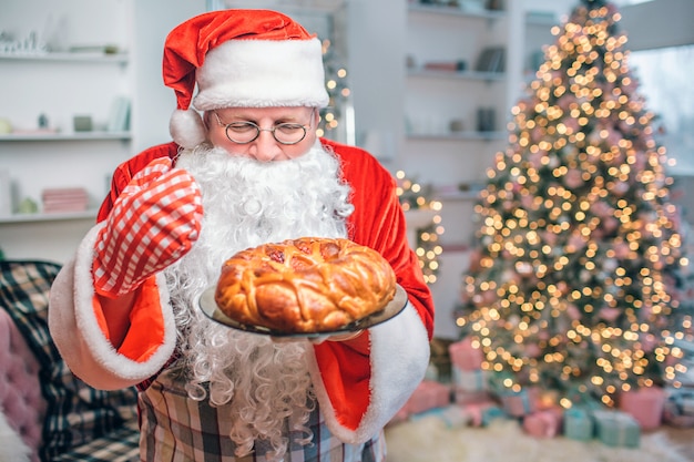 Świeże I Smaczne Ciasto Jest W Rękach świętego Mikołaja. Patrzy Na To I Pachnie. Za Nim Jest Choinka.