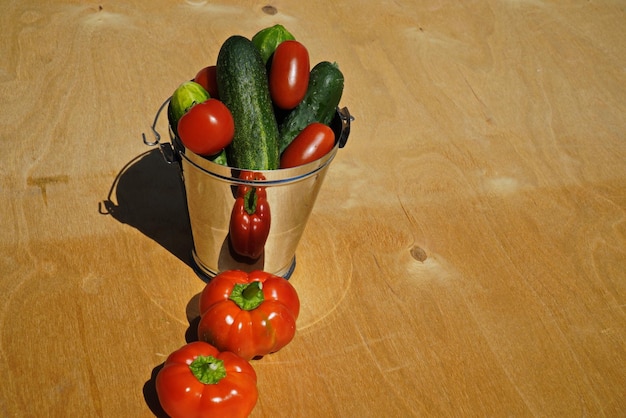 Świeże ogórki i pomidory w ozdobnym wiaderku obok dwóch czerwonych papryczek na naturalnym tle ze sklejki