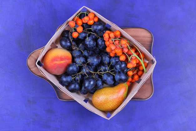 Świeże owoce na wegańskie zbliżenie Gruszka brzoskwinia jarzębina i ciemne winogrona w drewnianym koszu na drewnianej desce na niebieskim tle