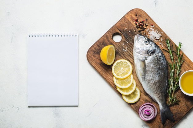 Zdjęcie Świeże surowe ryby dorado na rustykalnej drewnianej desce do krojenia z przyprawami i notatnikiem do przepisu lub menu. widok z góry na białym tle.
