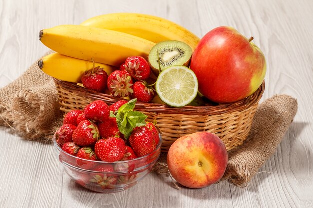 Świeże truskawki w szklanej misce zwieńczone liśćmi mięty, nektaryną, limonką, kiwi, bananem, jabłkiem w wiklinowym koszu na drewnianym stole