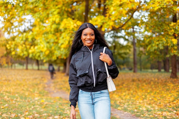 Świeży portret pięknej szczęśliwej młodej czarnej dziewczyny z uśmiechem w modnej casualowej kurtce i dżinsach z torebką spaceruje po jesiennym parku ze złotymi jesiennymi liśćmi