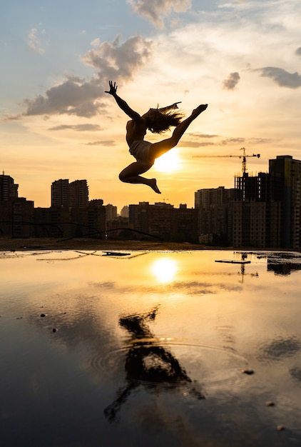 Sylwetka kobiece tancerki elastyczne skoki podczas zachodu słońca na tle miasta z odbiciem w wodzie